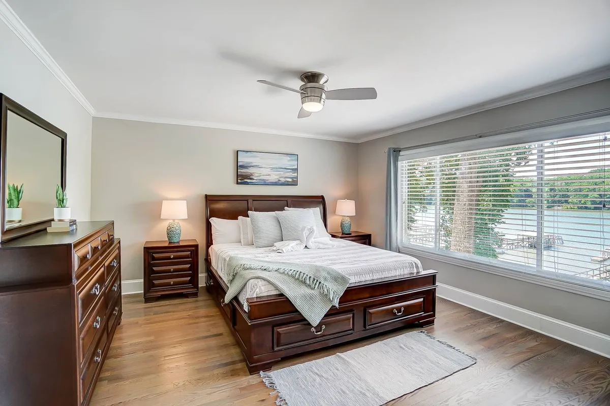 This image shows a bedroom featuring a large window that offers a scenic view of the outdoors. The bed is invitingly made, and the room is decorated with peaceful and natural tones, with decorative elements adding charm. The window view hints at a natural landscape, suggesting the presence of greenery or parkland just outside.