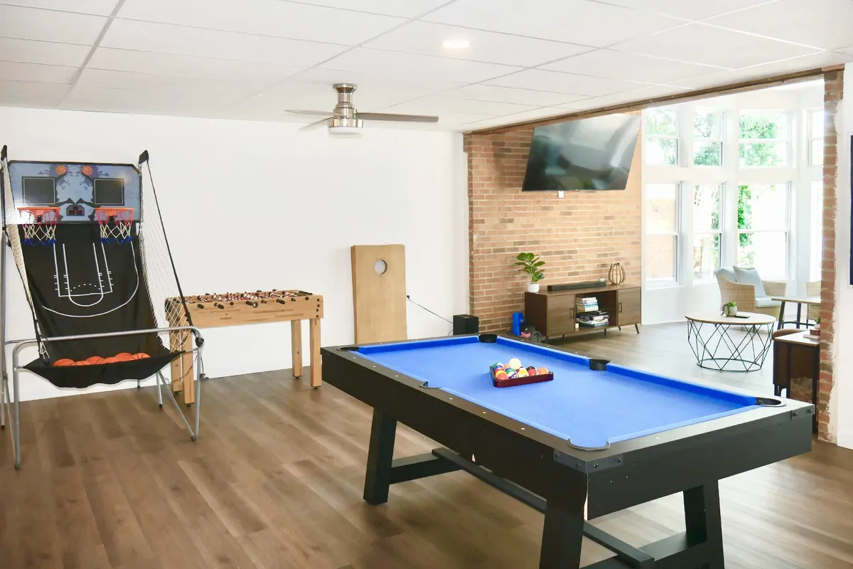 This image showcases a vibrant recreation room featuring a pool table, foosball table, basketball arcade game, and large windows that allow plenty of natural light and offer scenic views. A comfortable couch invites guests to relax and socialize after playing games.