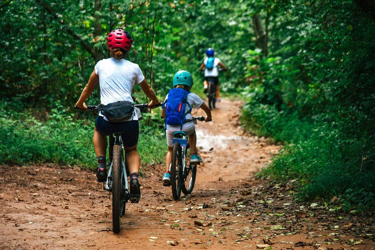 A joyful exterior scene showing bicycles parked along a forest path, with children and families actively exploring the wooded area. The mood is energetic and playful, emphasizing discovery and family bonding in a natural setting.