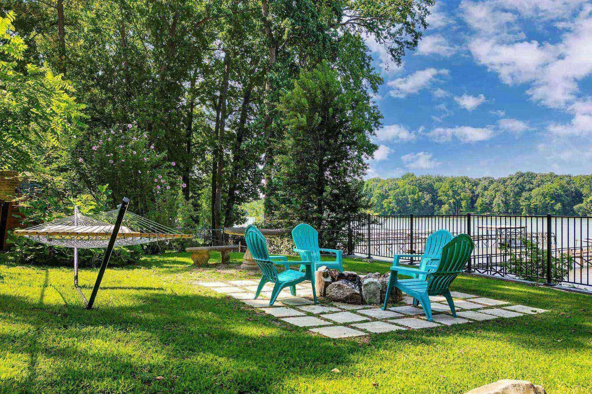 A cozy outdoor setup with turquoise chairs arranged around a fire pit on a manicured lawn, overlooking the river or lake. The fire pit area is inviting, suggesting warm evenings spent under the stars with friends or family. The water view in the background enhances the tranquil and communal vibe of the space.