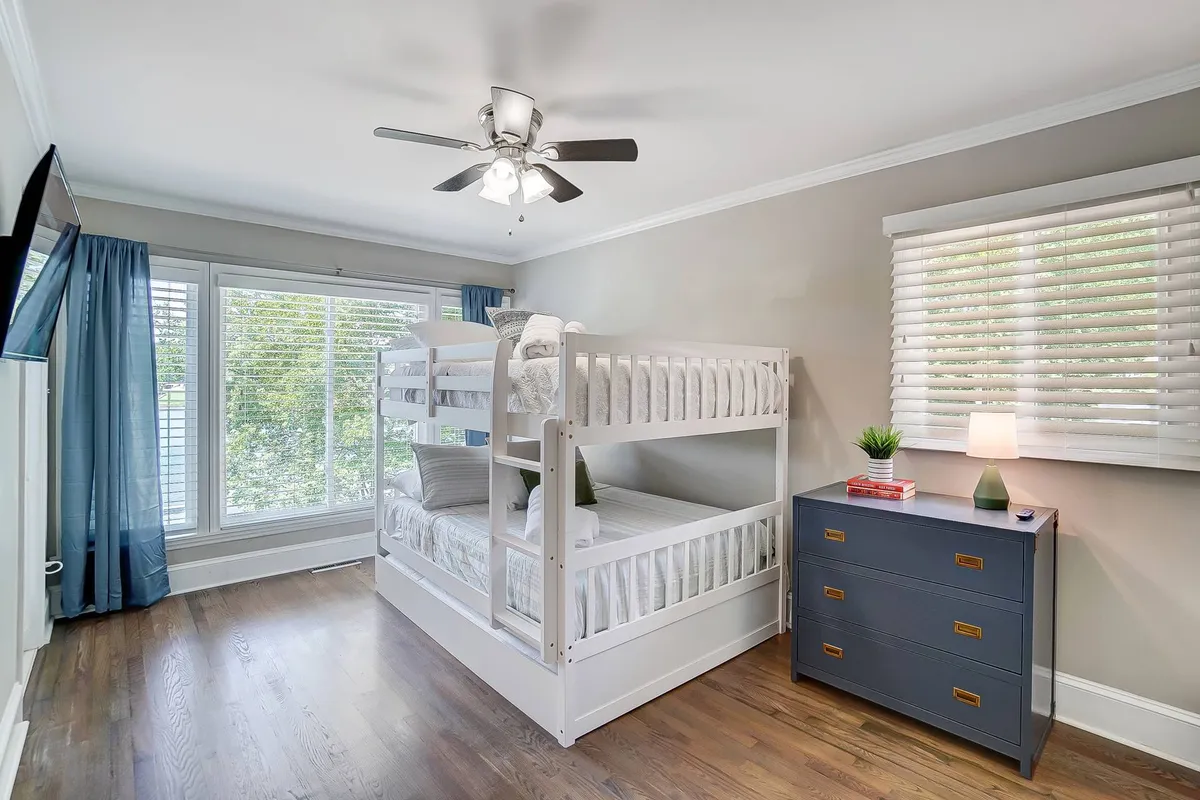 A cozy room with a bunk bed set next to a large window filled with bright, natural light. The dresser and lamp add a homey atmosphere, while the window’s size and placement suggest a connection to the outdoors. The playful arrangement of the bunk bed highlights a family-friendly environment ideal for children and group stays.