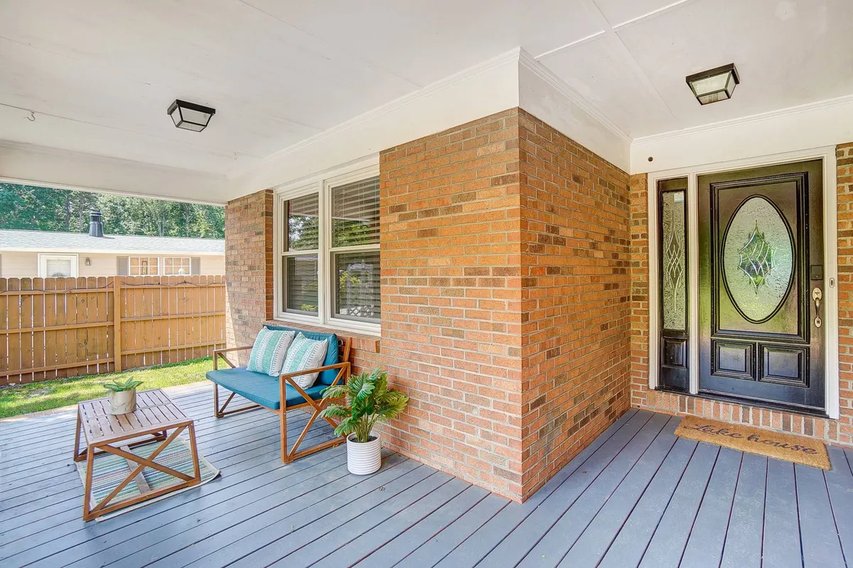 A close-up of the front door and outdoor seating area, accented by a well-kept planter and cheerful doormat. The scene suggests hospitality and a sunny spot for morning coffee or greeting friends before heading out.