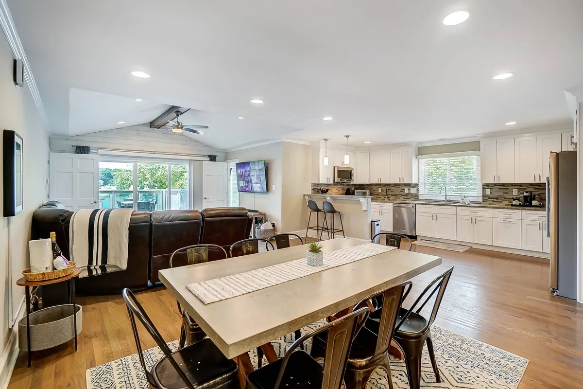 A spacious, open-plan living area that integrates a dining table, kitchen island, large windows, and a comfortable seating arrangement. Natural light floods the space, highlighting the cozy yet modern interior where guests can gather, dine, and relax together.